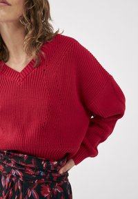 Czerwony sweter z dzianiny z dekoltem w serek, z teksturowanym ściągaczem i ozdobnymi wzorami. W zestawie z czarną spódnicą ozdobioną kwiatowymi nadrukami.