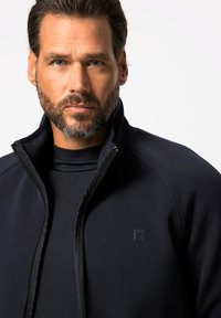 Chaqueta con cremallera de color azul marino oscuro y cuello alto, de textura lisa. Presenta un logotipo en el pecho y un diseño ajustado.