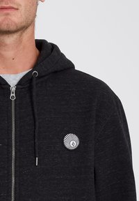 Black zip-up hoodie made of textured fabric with a large round logo on the left chest, featuring sunburst detailing in white.