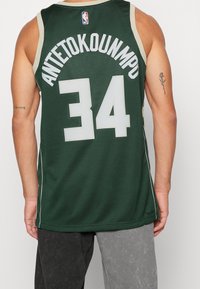 La maglia NBA verde presenta il nome "Antetokounmpo" e il numero "34" in bianco. Il design include un bordo chiaro a contrasto e cuciture laterali.