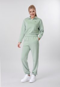 Γυναίκα όρθια με ανοιχτοπράσινο φόρμα και λευκά αθλητικά παπούτσια, με το χέρι στην τσέπη, χαμογελαστή μπροστά σε ένα απλό ανοιχτό γκρι φόντο.