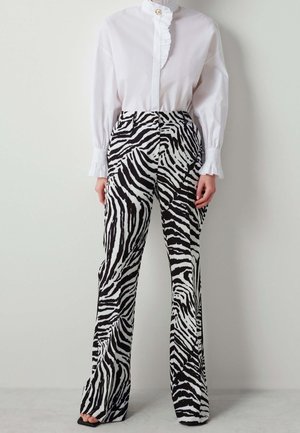 Kvinde iført sort-hvide zebrastribede bukser kombineret med en hvid bluse med flæset krave og manchetter, stående mod en ensfarvet baggrund.