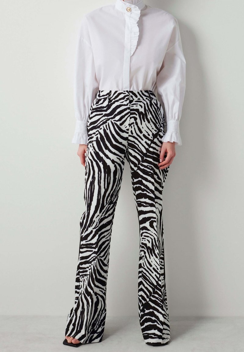 Femme portant un pantalon imprimé zèbre noir et blanc associé à une blouse blanche avec col et poignets à volants, debout devant un fond uni.