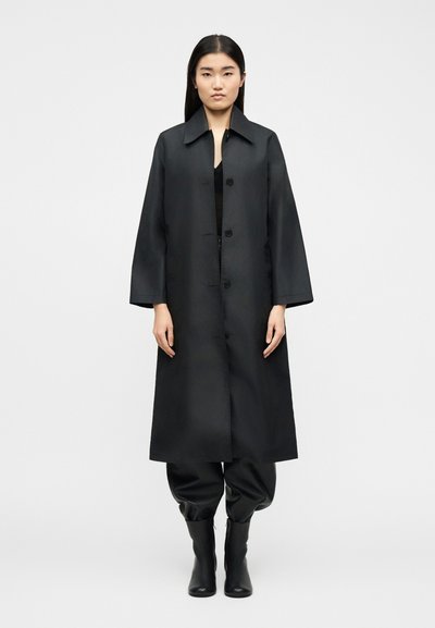 Femme debout face à l'avant, portant un long manteau noir boutonné, un pantalon noir ample et des bottines noires, sur un fond uni.