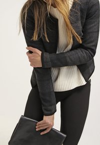 Blazer a righe nere e grigie sopra un maglione bianco a coste, abbinato a leggings neri, mentre tiene una pochette in pelle nera.