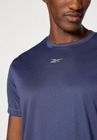 Navy blue athletic t-shirt with short sleeves and a crew neckline, featuring a subtle logo in light gray on the upper chest area. Smooth fabric.