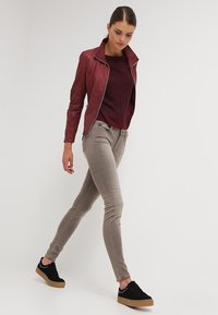 Giacca di pelle rossa, maglione tarocca bordeaux, jeans slim-fit grigi chiari, sneakers nere con suola color sabbia e borchie, posa in cammino su sfondo neutro.