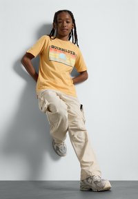 Camiseta amarilla de manga corta con el logo de "Quiksilver" y gráfico de atardecer; combinada con pantalones cargo beige y zapatillas grises.