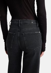 Zwarte spijkerbroek met hoge tailleband, vijf zakken en een klein logolabel aan de achterkant. De stof heeft een gladde, licht gestructureerde afwerking.