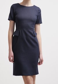 Mörkblå klänning med kort ärm, texturerad tyg, figursydd design och sidopaneler i peplum. Rund hals, knäskort längd.