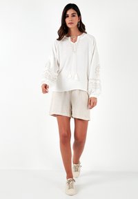 White long-sleeve top with floral embroidery and tie detail at the neckline, paired with beige linen shorts and light-colored sneakers.