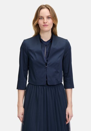 Giacca cropped blu navy realizzata in tessuto liscio, con maniche a tre quarti e chiusura con un unico bottone, indossata sopra un vestito plissettato.