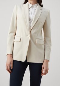 Blazer de corte ajustado en color beige con cierre de un solo botón, solapas en pico y dos bolsillos frontales. Combinado con una camisa blanca de cuello.