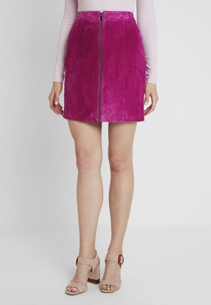 Kobieta ubrana w jasnoróżową mini spódnicę zamszową z przednim zamkiem błyskawicznym, w połączeniu z beżowymi sandałami na obcasie typu słupek z odkrytymi palcami oraz jasnoróżową, długorękawową bluzką.