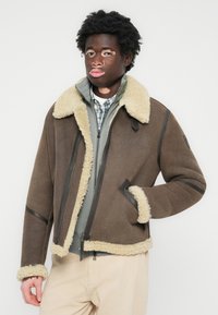 Chaqueta de shearling marrón con un cuello de piel beige, cierre con cremallera en la parte delantera y acentos oscuros en contraste. Llevada sobre una capa gris acolchada y una camisa de cuadros.
