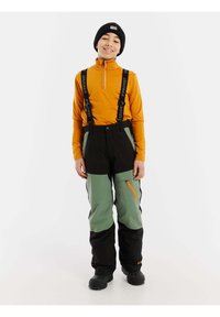 Maglia arancione a maniche lunghe con cerniera, pantaloni da sci verdi e neri con bretelle regolabili, vita elastica e tasca laterale con cerniera. Stivali neri.