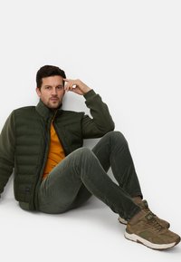 Grøn polstret vest over en grøn fleece, kombineret med en orange skjorte, grønne bukser og brune sneakers. Glat stof med syede mønstre.