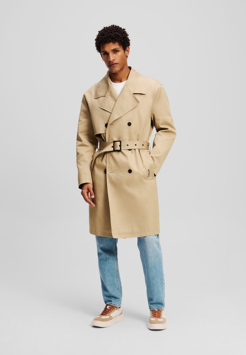 Trench beige con un diseño de doble botonadura, amplios solapas, cintura ceñida con cinturón y parte delantera abotonada. Combinado con jeans de color azul claro y zapatillas.