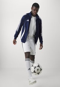 Marine Adidas jakke med hvite striper, hvit t-skjorte, hvite shorts og fotballsokker med svart stripe. Fotball ved føttene.