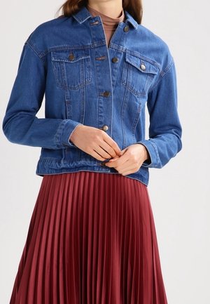Woman wearing a blue denim jacket with button details over a brown turtleneck and a high-waisted, pleated red skirt.