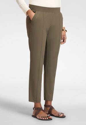 Frau trägt olivgrüne gerade geschnittene Hose mit Seitentaschen, ein weißes Oberteil, ein goldenes Armband und braune Sandalen mit offenem Zehenbereich und Knöchelriemchen.