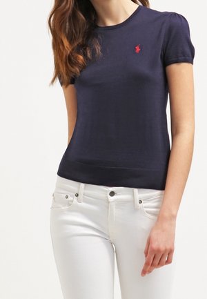 Femme portant un haut ajusté à manches courtes bleu marine avec un petit logo rouge sur la poitrine et un jean blanc, debout devant un fond uni.