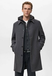 Abrigo de lana gris con cuello, que cuenta con cierre de botones, bolsillos laterales y un chaleco gris en capas debajo. Combinado con jeans oscuros.
