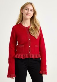 Rode gebreide cardigan met een gekruide zoom, sluiting met parelknopen en twee voorzakken. Lange mouwen hebben een gerimpelde rand. Zacht materiaal.