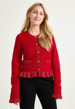 Červený pletený cardigan s volánkovým lemem, uzávěrem na perlové knoflíky a dvěma předními kapsami. Dlouhé rukávy mají zakončení s volánkem. Měkká textura.