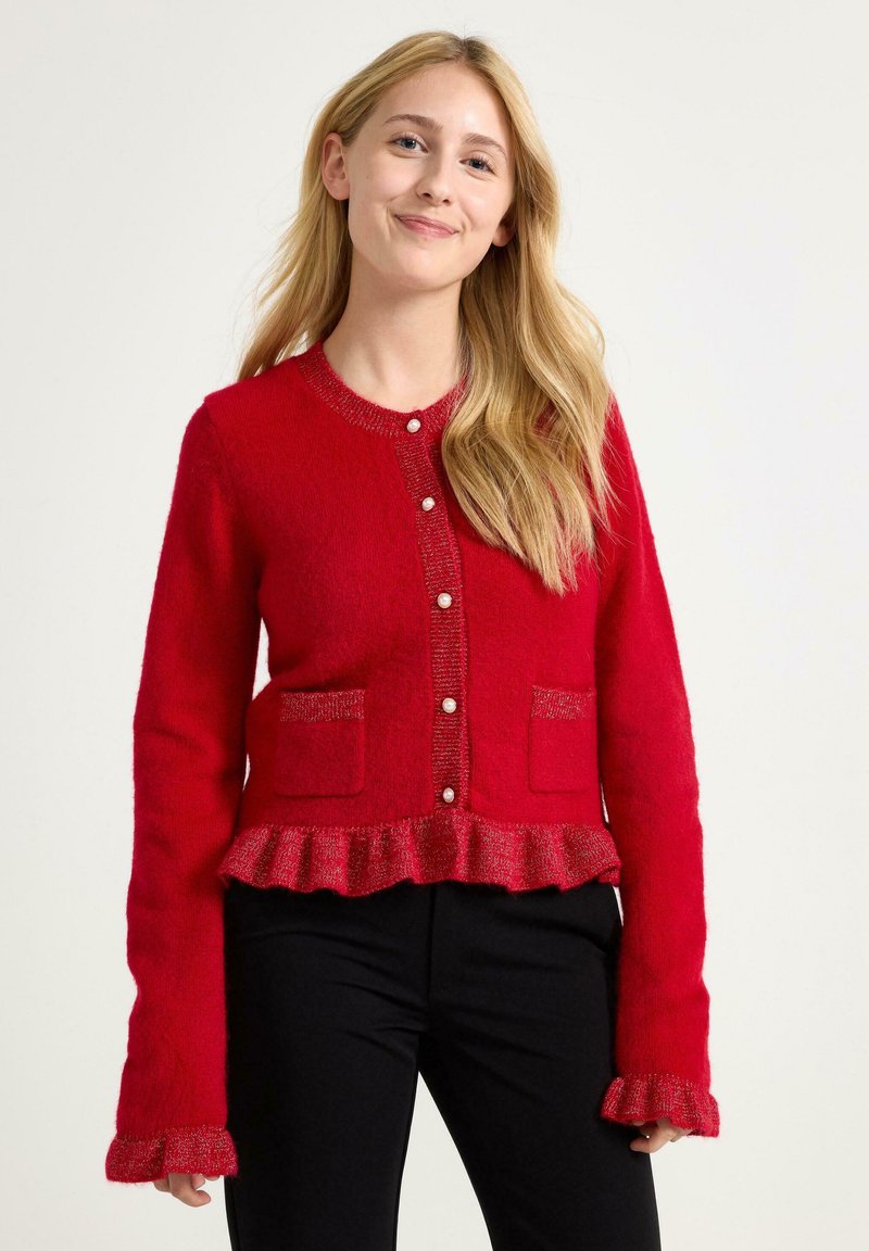 Red knitted cardigan with a ruffled hem, pearl button closure, and two front pockets. Long sleeves feature a frilled edge. Soft texture.