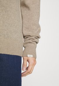 Beżowy sweter z dzianiny z prążkowanymi mankietami, z wszywaną metką przy brzegu. Miękka struktura, luźny krój, noszony na ciemnych dżinsach.