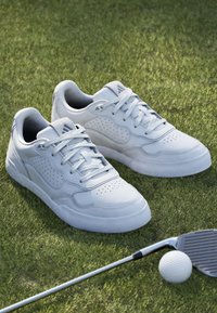 Vita lädersneakers med perforerade detaljer, platta snören och en slät tådel, placerade på gräs med en golfklubba och boll i närheten.