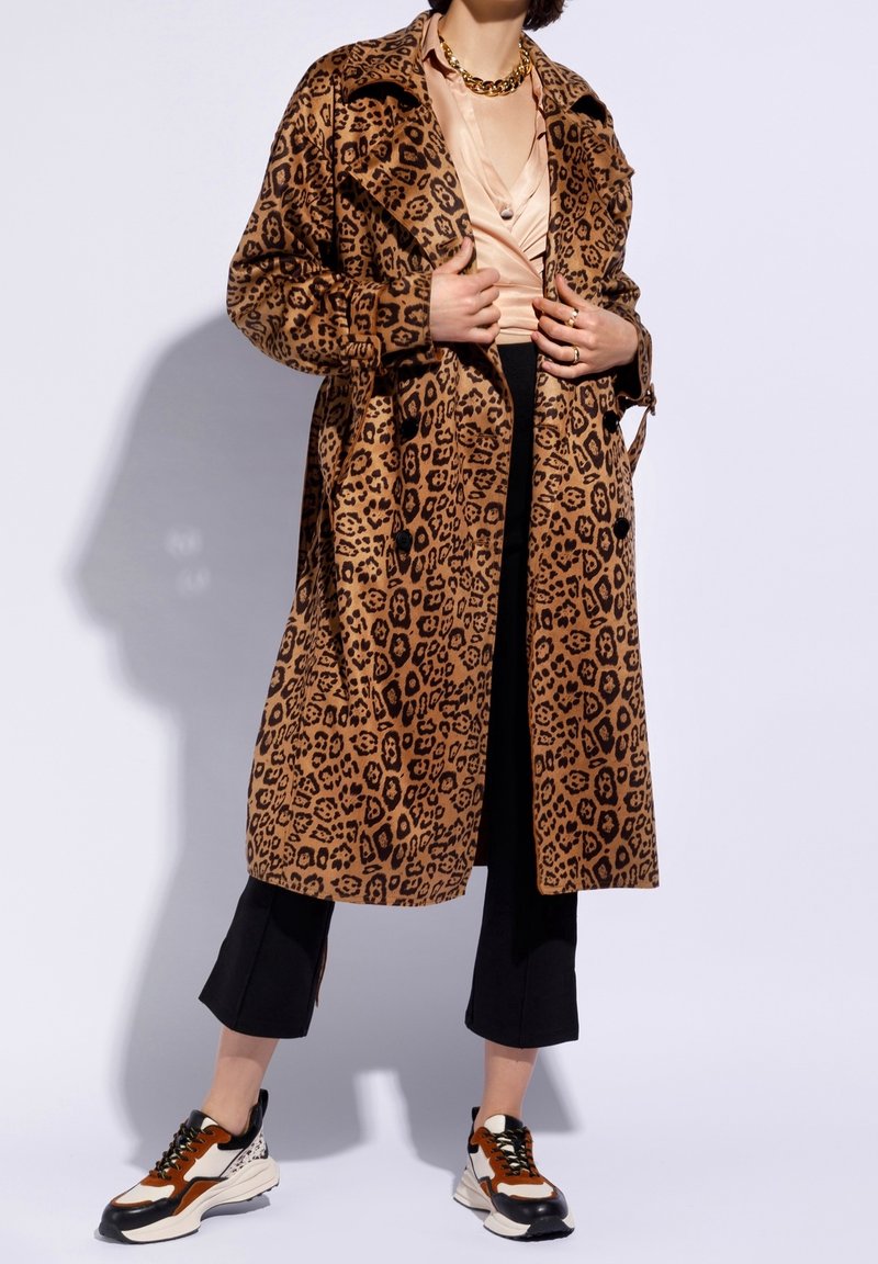 Personne portant un long manteau à imprimé léopard, un chemisier beige, un pantalon court noir, des baskets épaisses et un collier chaîne doré, debout devant un fond blanc.