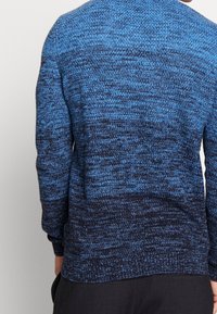 Pull en tricot bleu et bleu foncé avec un motif texturé en dégradé. Les manches longues et les poignets côtelés ajoutent du détail au design.