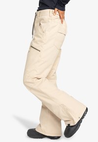 Beige cargo pants with a relaxed fit, featuring multiple pockets, zippers, and a slight flare at the leg. Made from durable fabric.