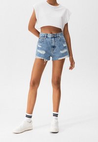 Persona vistiendo pantalones cortos de mezclilla azul desgastados, una blusa blanca sin mangas y corta, zapatillas blancas y calcetines blancos con rayas negras, de pie contra un fondo blanco.