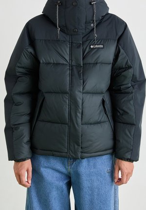 Chaqueta acolchada negra con capucha, cremallera frontal y bolsillos laterales, llevada con vaqueros azules. Logotipo de Columbia en el pecho.