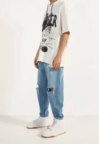 Kortärmad, krämfärgad hoodie med svart grafisk tryck; ihop med ljusblå slitna jeans och vita hög- topp sneakers.