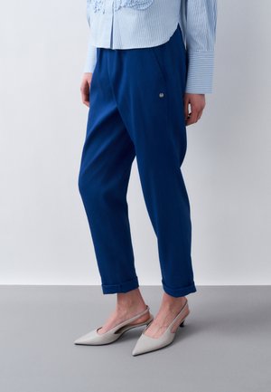 Γυναίκα με μπλε παντελόνια με μανσέτες, ριγέ πουκάμισο με κεντήματα και ανοιχτά ψηλοτάκουνα παπούτσια σε ανοιχτό γκρι, στέκεται σε γκρι πάτωμα.