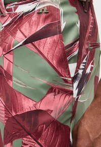 Suur täiuslikkus rohelistest lühikestest pükstest, millel on suur punase ja roosa troopilise lehe muster, metallist logo tasku lähedal ja käsi riideeseme kõrval lõõgastumas.