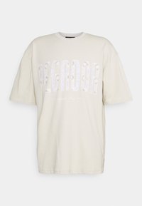 Beige oversized T-shirt med korta ärmar och stor mönstrad text "PEGADOR" över bröstet, med mindre text nedanför som lyder "Atlantic Peace."