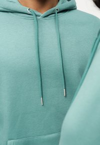 Πράσινη φούτερ μπλούζα με κορδόνι, που διαθέτει ασημί άγκιστρα, μαλακό ύφασμα και μπροστινή τσέπη. Κοντινή λήψη που υπογραμμίζει την υφή και το σχέδιο.