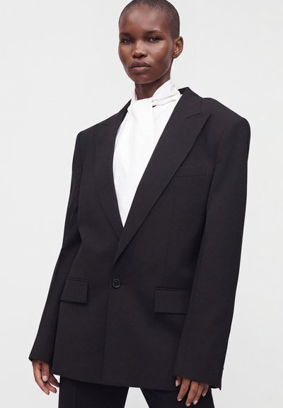 Femme portant un blazer noir oversize avec des poches à rabat et un seul bouton sur un chemisier blanc à col montant.