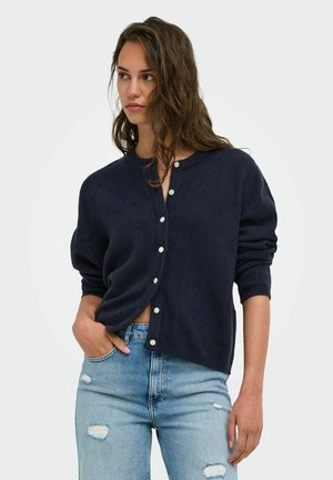 Femme aux longs cheveux bruns portant un pull bleu marine foncé boutonné et un jean bleu clair déchiré, regardant vers sa gauche avec une main dans la poche.