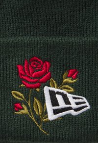 Gr�üner Strickbeanie mit einer roten Rose und goldenen Blättern, die neben einem weißen und schwarzen Logodetail gestickt sind. Weiche Textur, engmaschiges Design.