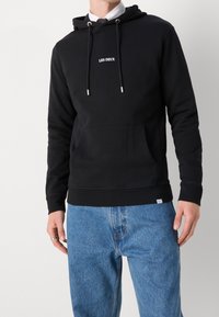 Czarna bluza z kapturem, z przednią kieszenią, regulowanymi sznurkami do ściągania i wyszywanym logo "LES DEUX". Ściągacze na rękawach i u dołu, w połączeniu z jasnoniebieskimi dżinsami.