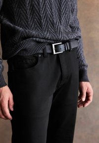 Sötét szürke kötött pulóver halszálkás mintával, fekete nadrággal párosítva, amelyet egy matt fekete öv rögzít, ezüst csattal.