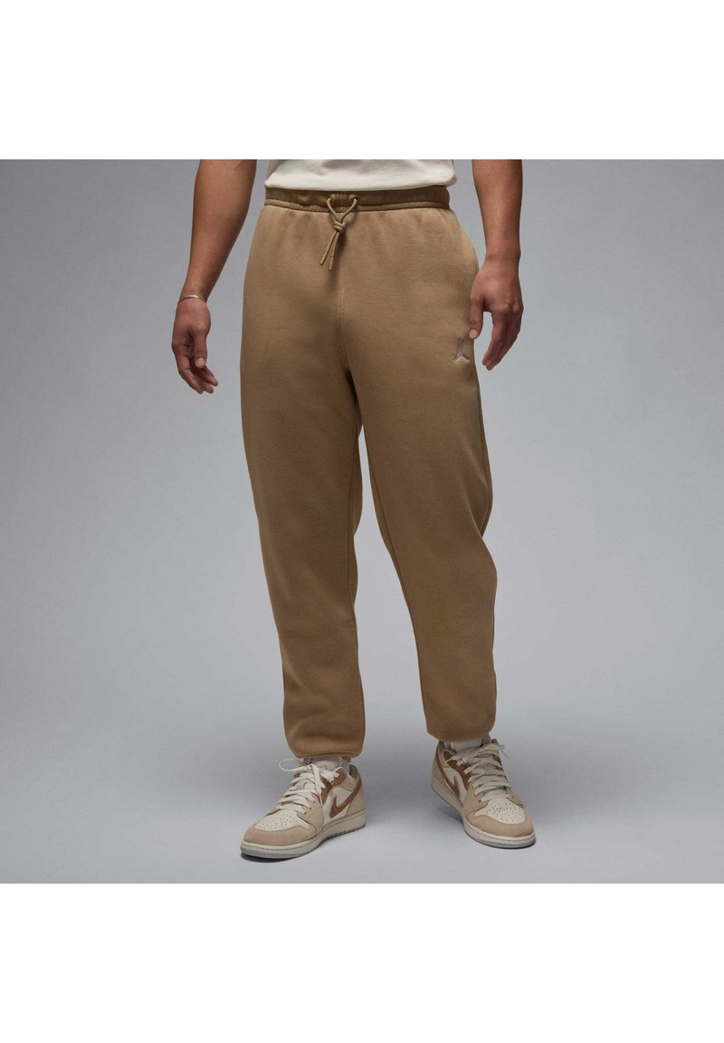Brown cotton joggers with a drawstring waist and side pockets. Features a small logo on the left thigh. Paired with beige sneakers.