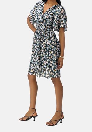 Femme portant une robe noire fleurie longueur genoux avec des manches volantées et des sandales noires à talons avec des lanières, debout sur un fond clair.