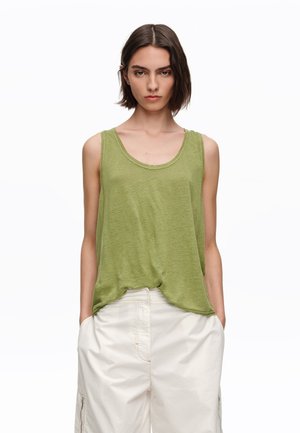 Kobieta z krótkimi brązowymi włosami ubrana w luźny zielony top bez rękawów i białe szerokie spodnie, z rękami w kieszeniach, zwrócona przodem.